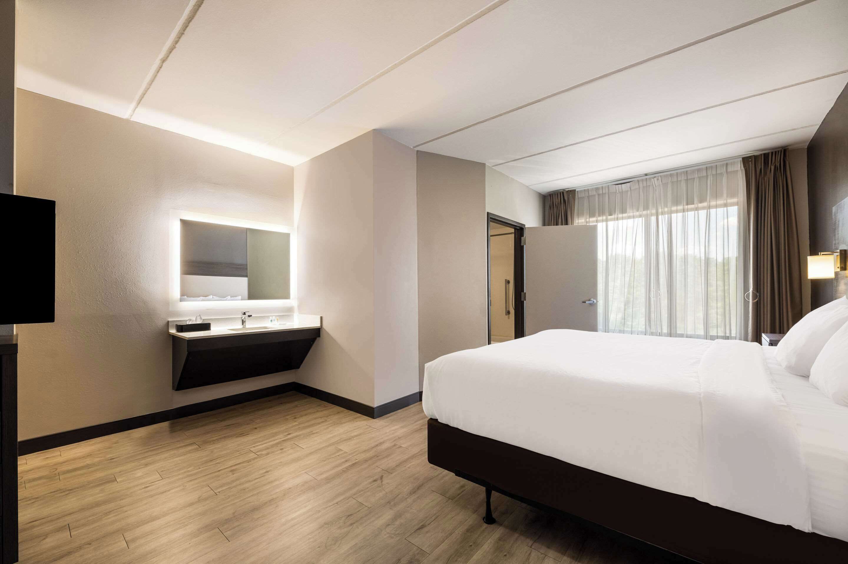 A hotel room with a large bed, a built-in vanity with a mirror, and a window with curtains.