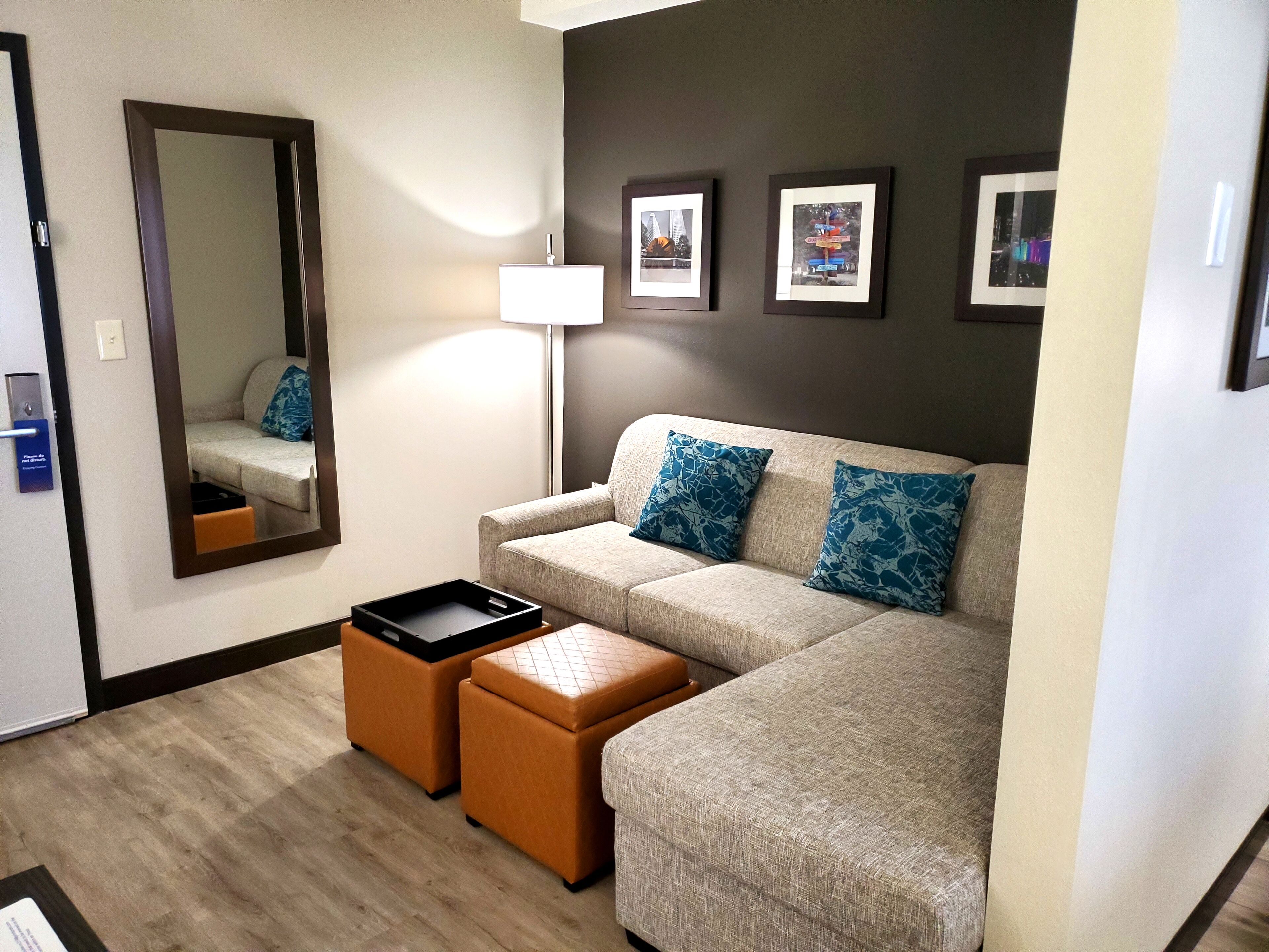 A modern hotel room with a sofa, ottomans, a mirror, and framed pictures on the wall.