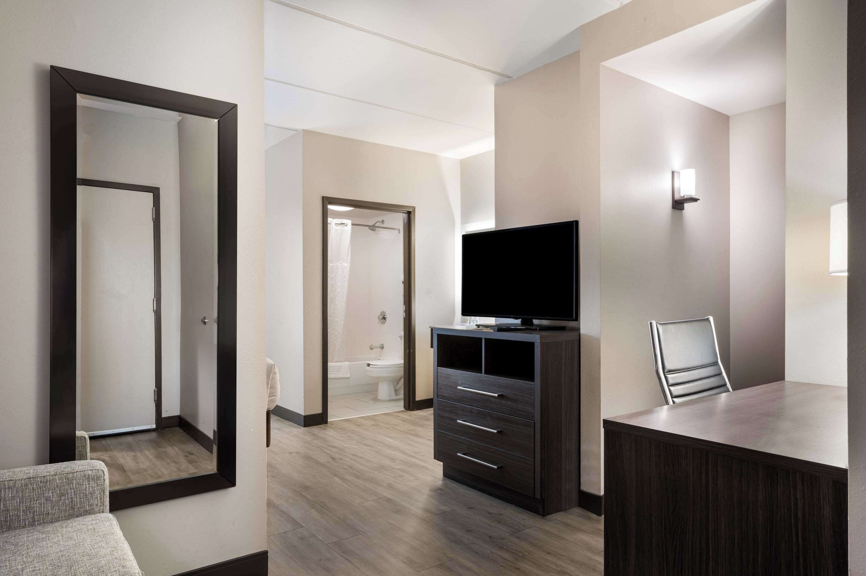 A modern hotel room with a large mirror, a flat-screen TV on a dark wooden cabinet, a small desk with a chair, and a shower area visible through an open door.