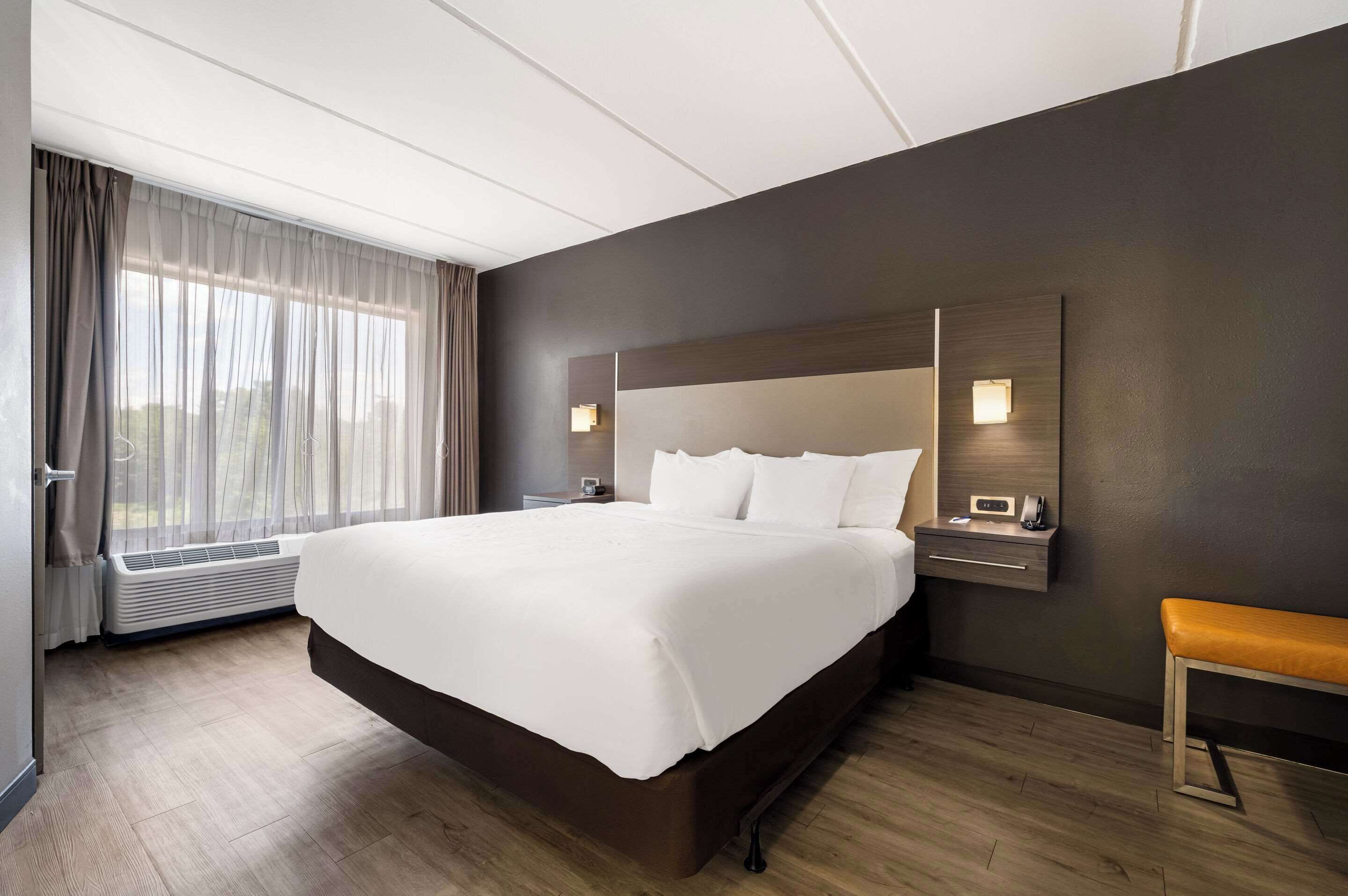 Een moderne hotelkamer met een groot bed, een nachtkastje met lamp, een bankje en een groot raam met gordijnen.