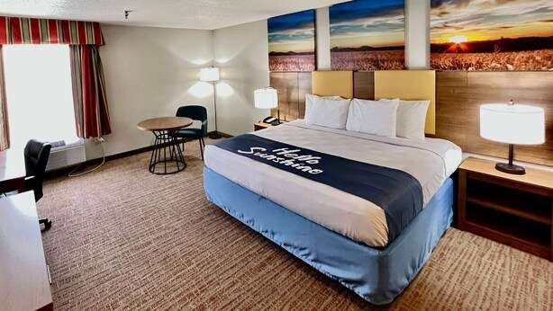 A hotel room with a large bed, two bedside lamps, a desk with a chair, and a view of a sunset through the window. - Days Inn by Wyndham Rayne/Crowley (Rayne)