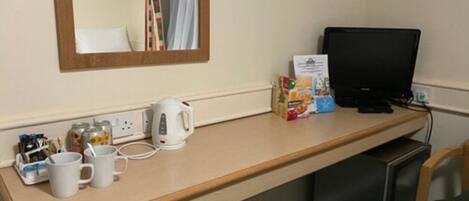 A hotel room with a wooden desk, a kettle, a television, and a chair.