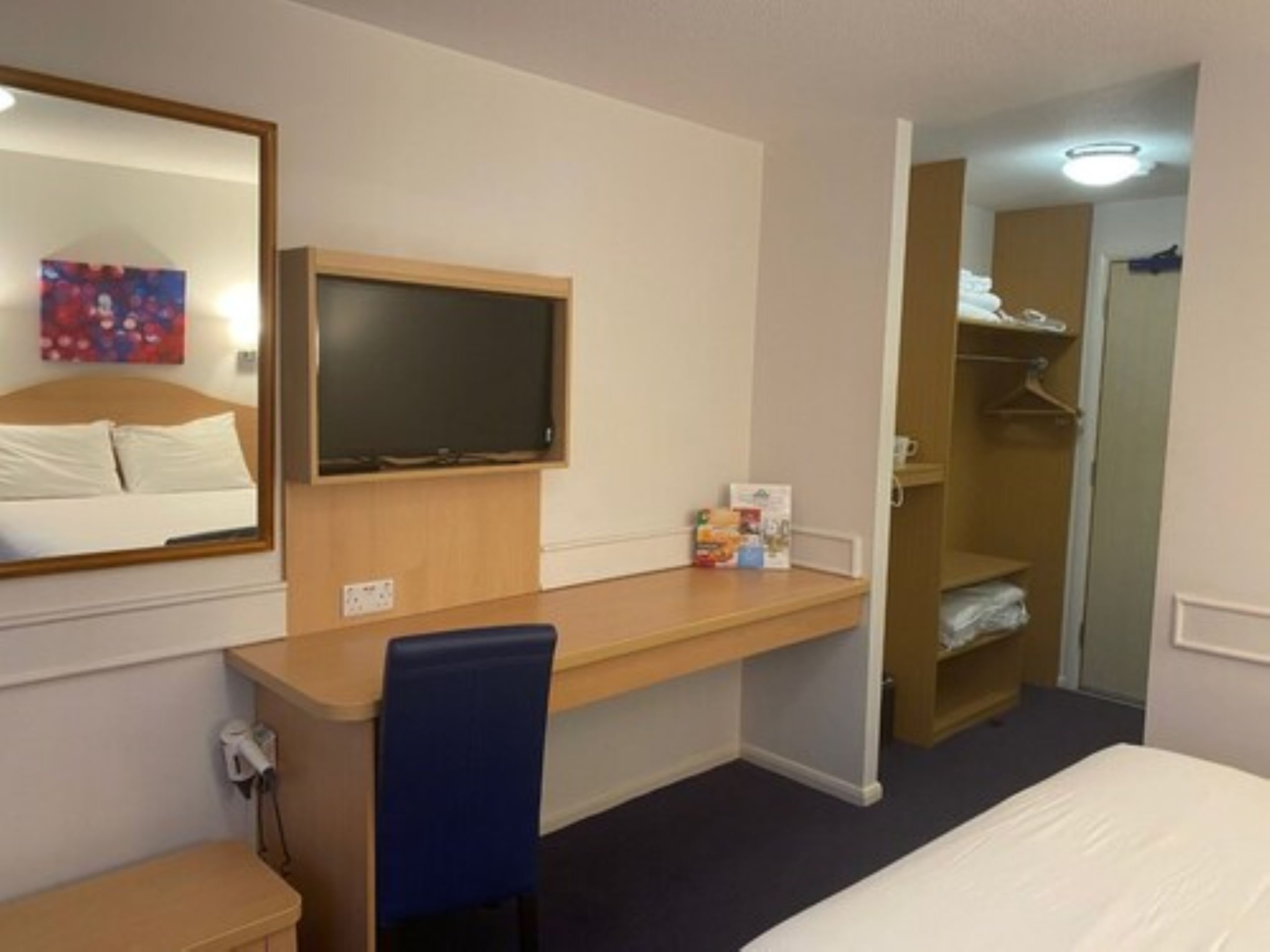 A hotel room with a bed, a desk with a chair, a television, a wardrobe, and a mirror.