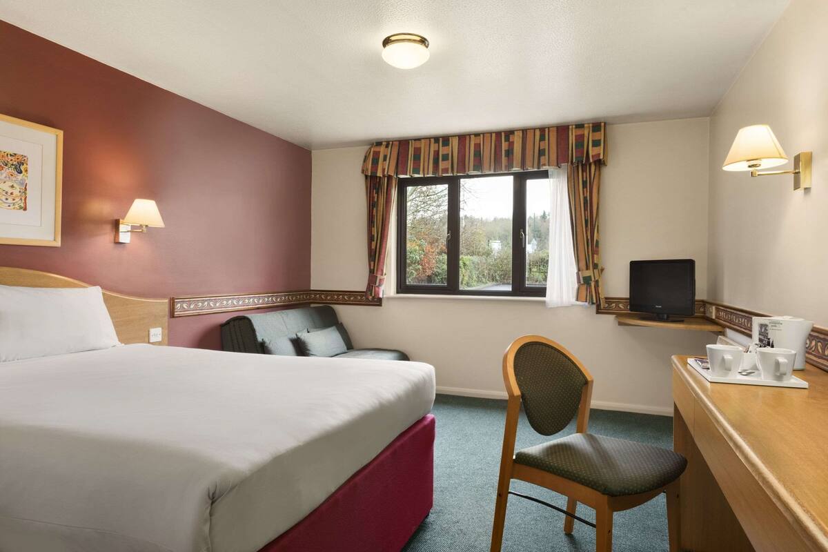 Habitación de hotel con cama, escritorio, silla, televisor y ventana con cortinas.