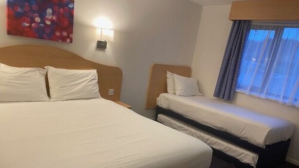 A hotel room with a large bed, a smaller bed, a nightstand, a lamp, a painting on the wall, and a window with curtains. - Days Inn by Wyndham Michaelwood M5 (Berkeley)