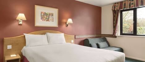 A hotel room with a large bed, two bedside lamps, a framed artwork above the bed, a chair, and a window with curtains.