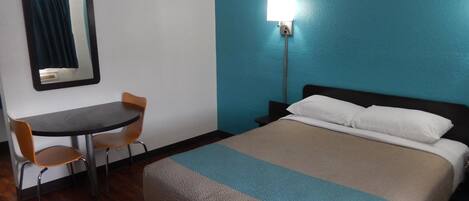 A hotel room with a bed, a desk, a chair, and a nightstand with a lamp.