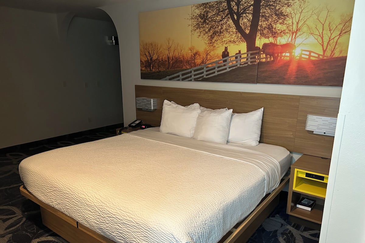 A hotel room with a large bed, a wooden headboard, a nightstand with a phone, a small table with a book, and a painting of a sunset scene on the wall.