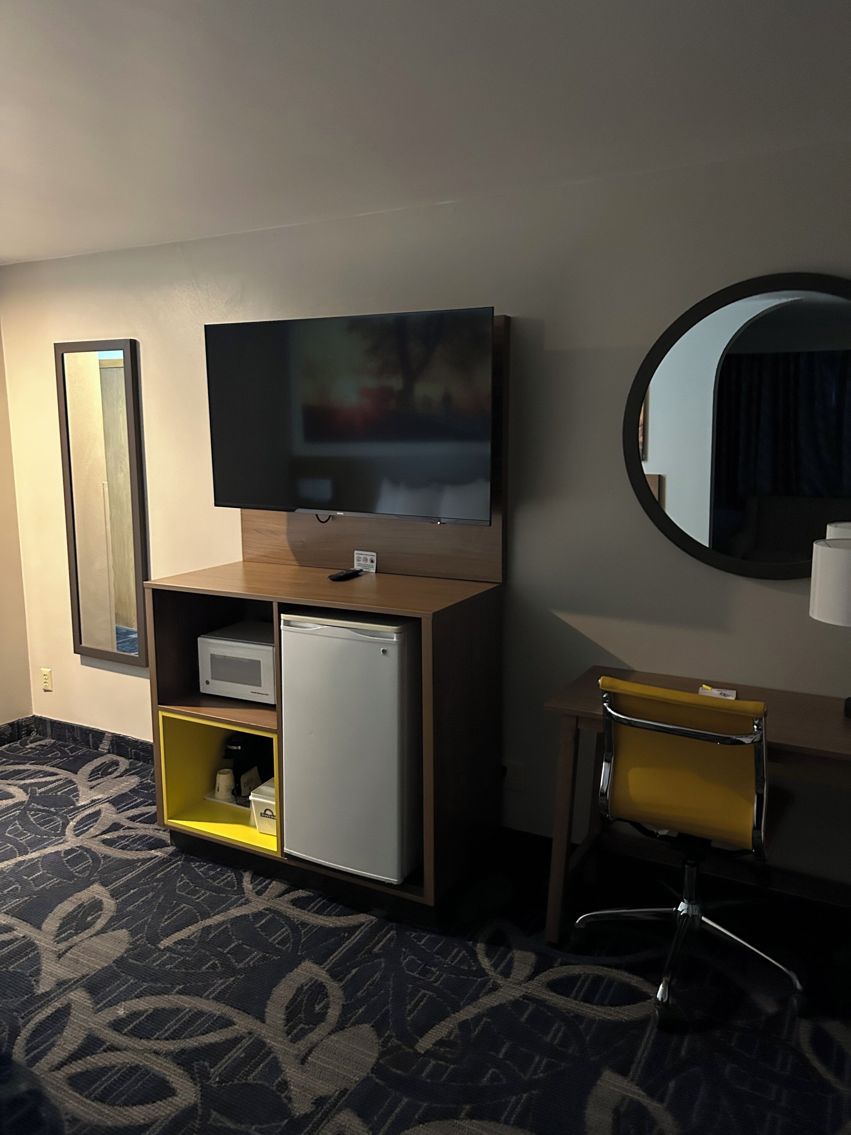 Habitación de hotel con televisor montado en un mueble de madera, un refrigerador pequeño, una silla amarilla y un espejo en la pared.