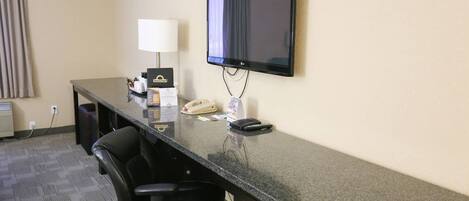 A hotel room with a desk, two chairs, a flat-screen TV, a lamp, a telephone, and a minibar.