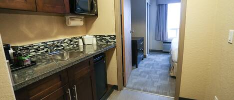 A compact kitchen with a microwave, sink, and dishwasher. A hallway leads to a bedroom with a window.