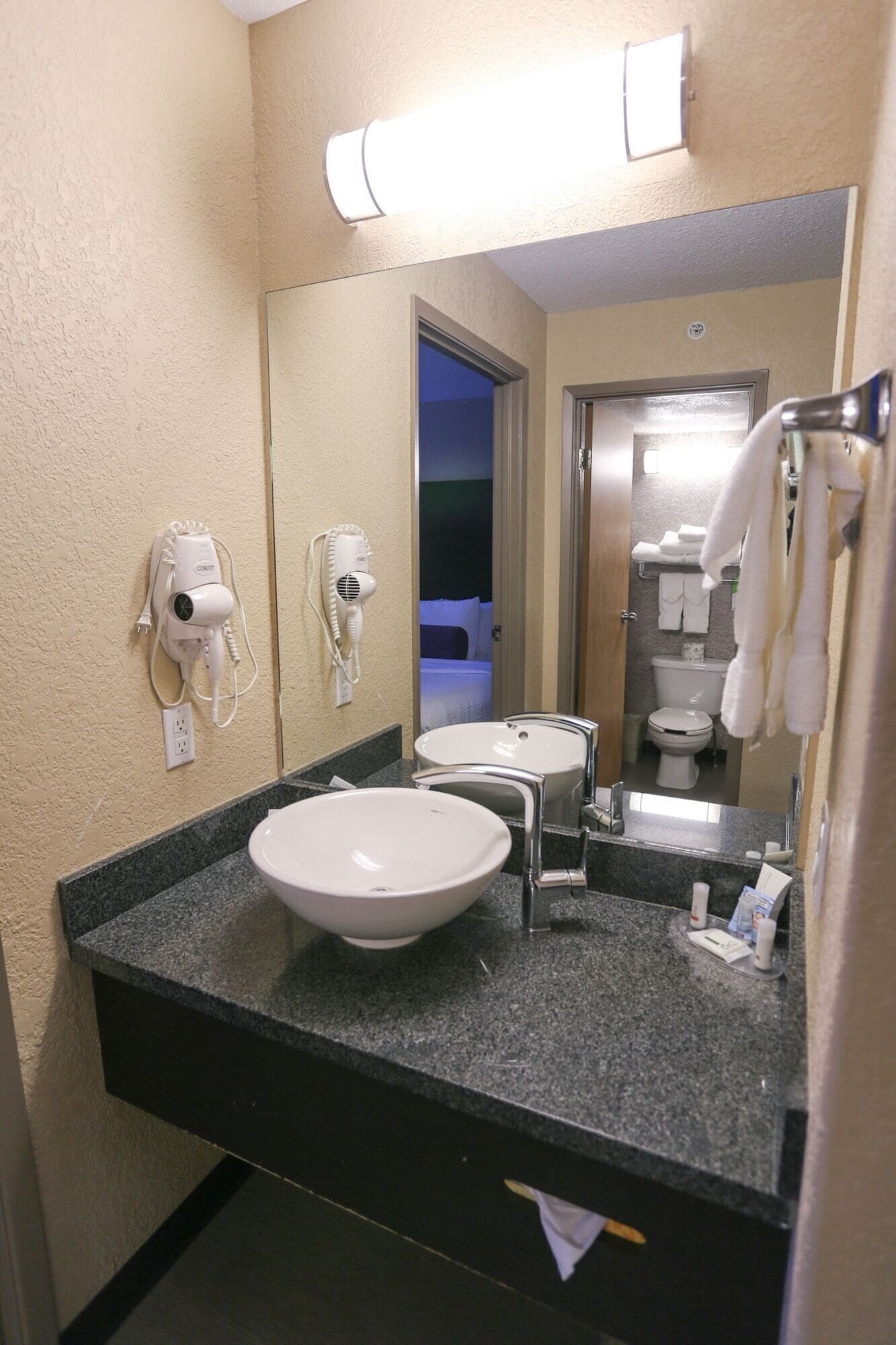 A hotel bathroom with a sink, two hair dryers, a mirror, and a toilet.