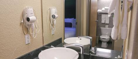 A hotel bathroom with a sink, two hair dryers, a mirror, and a toilet.
