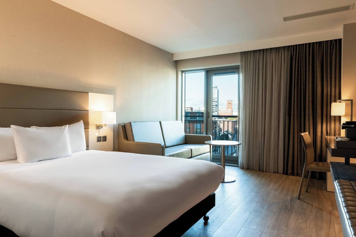 A hotel room with a large bed, a sofa, a small table, and a desk. There is a view of the city through the window.