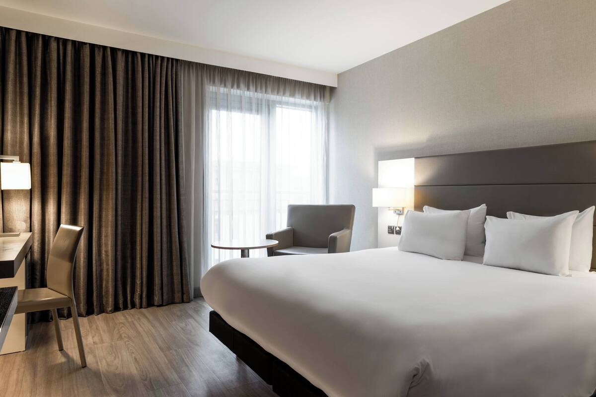 A hotel room with a large bed, a desk, a chair, a small table, and a window with curtains.