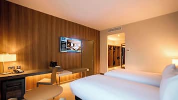 A hotel room with a wooden wall, a TV mounted on the wall, a desk with a chair, a bed with white bedding, and a minibar.