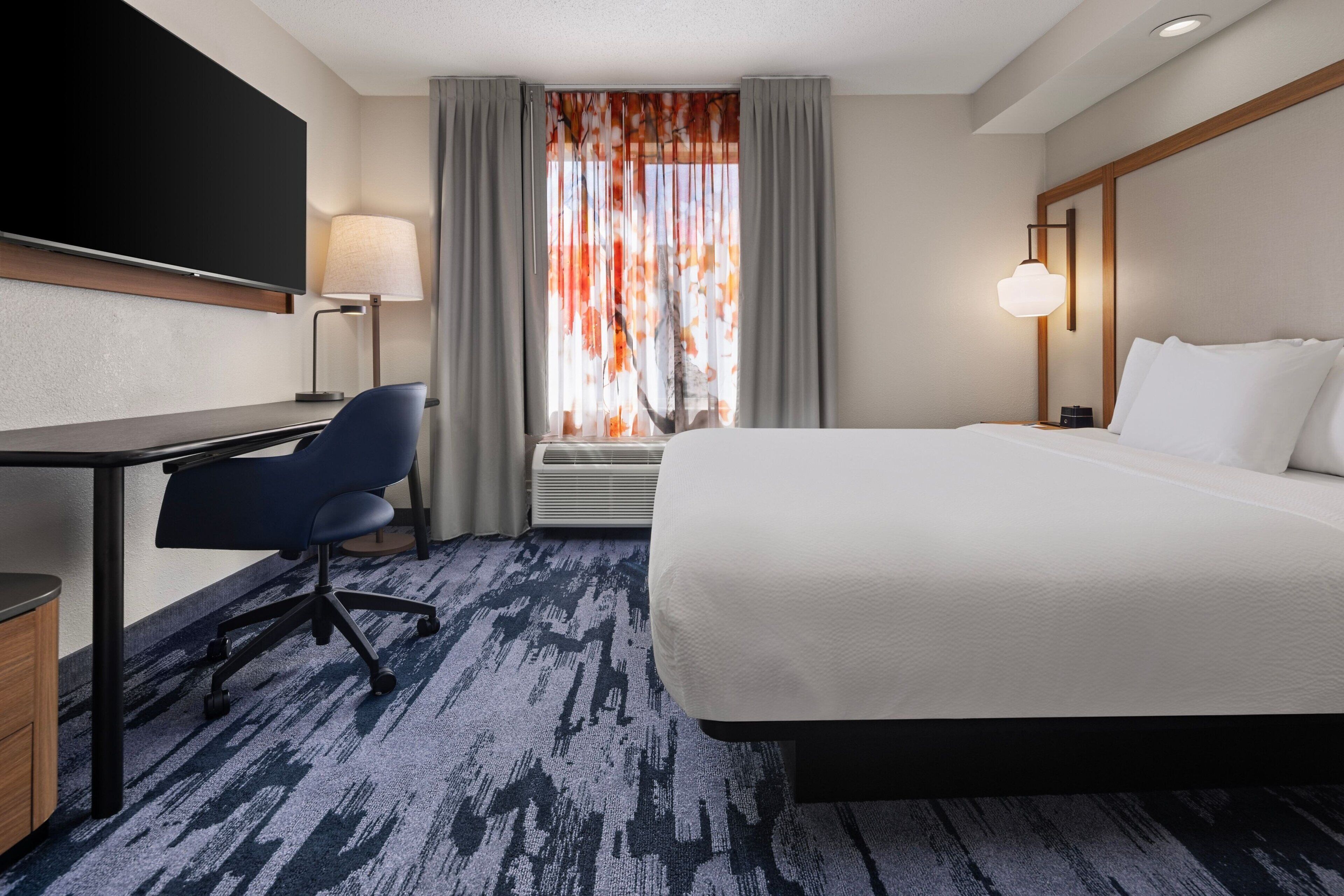 A hotel room with a large bed, a desk with a chair, a flat-screen TV, a lamp, and a window with curtains.