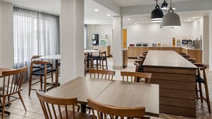 Un espace de salle à manger moderne avec des tables et des chaises en bois, une cuisine en arrière-plan et des luminaires suspendus.