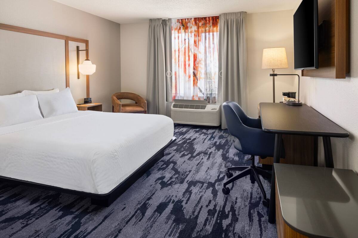 A hotel room with a large bed, a desk, a chair, a TV, and a window with curtains.