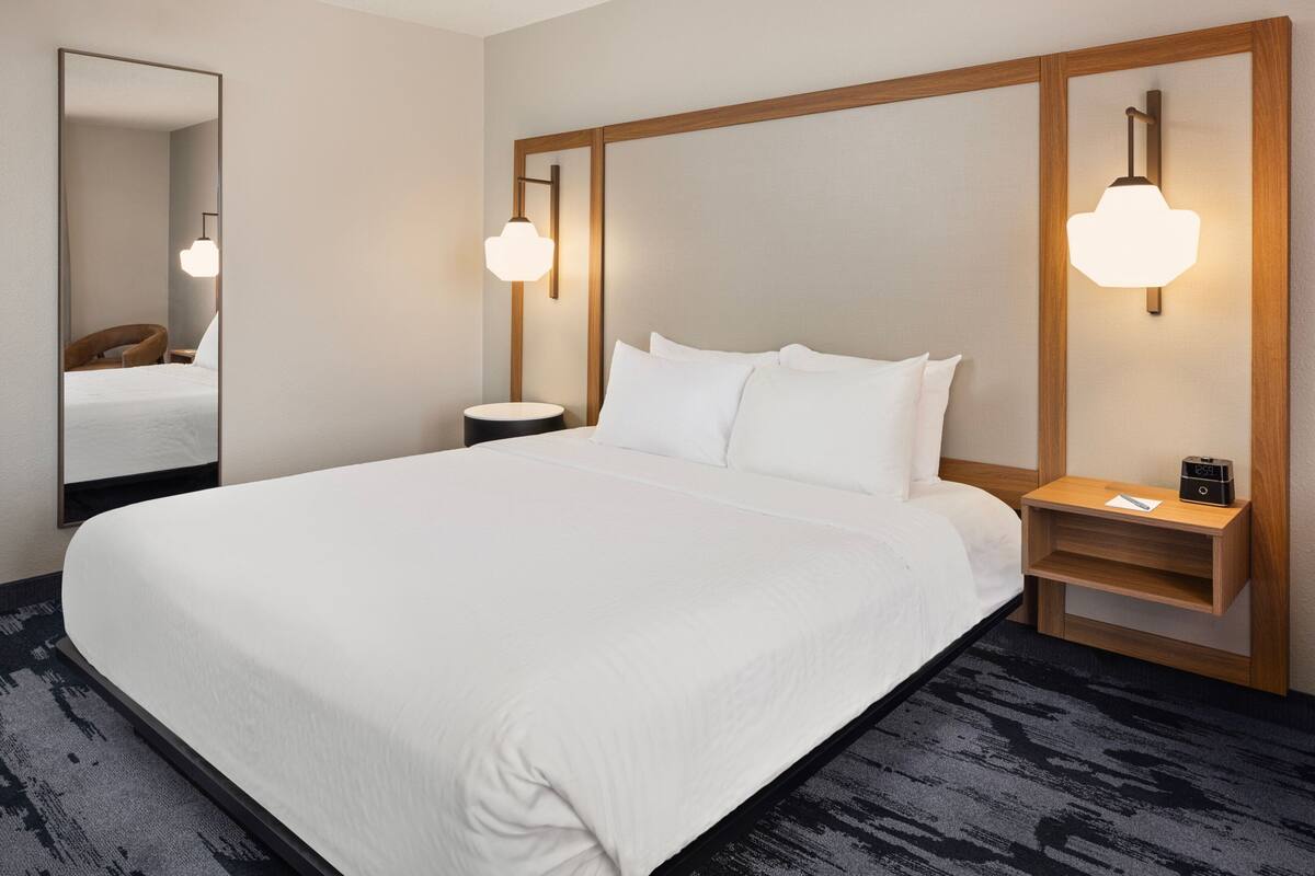 A hotel room with a large bed, a nightstand with a lamp, a mirror, and a wooden headboard.
