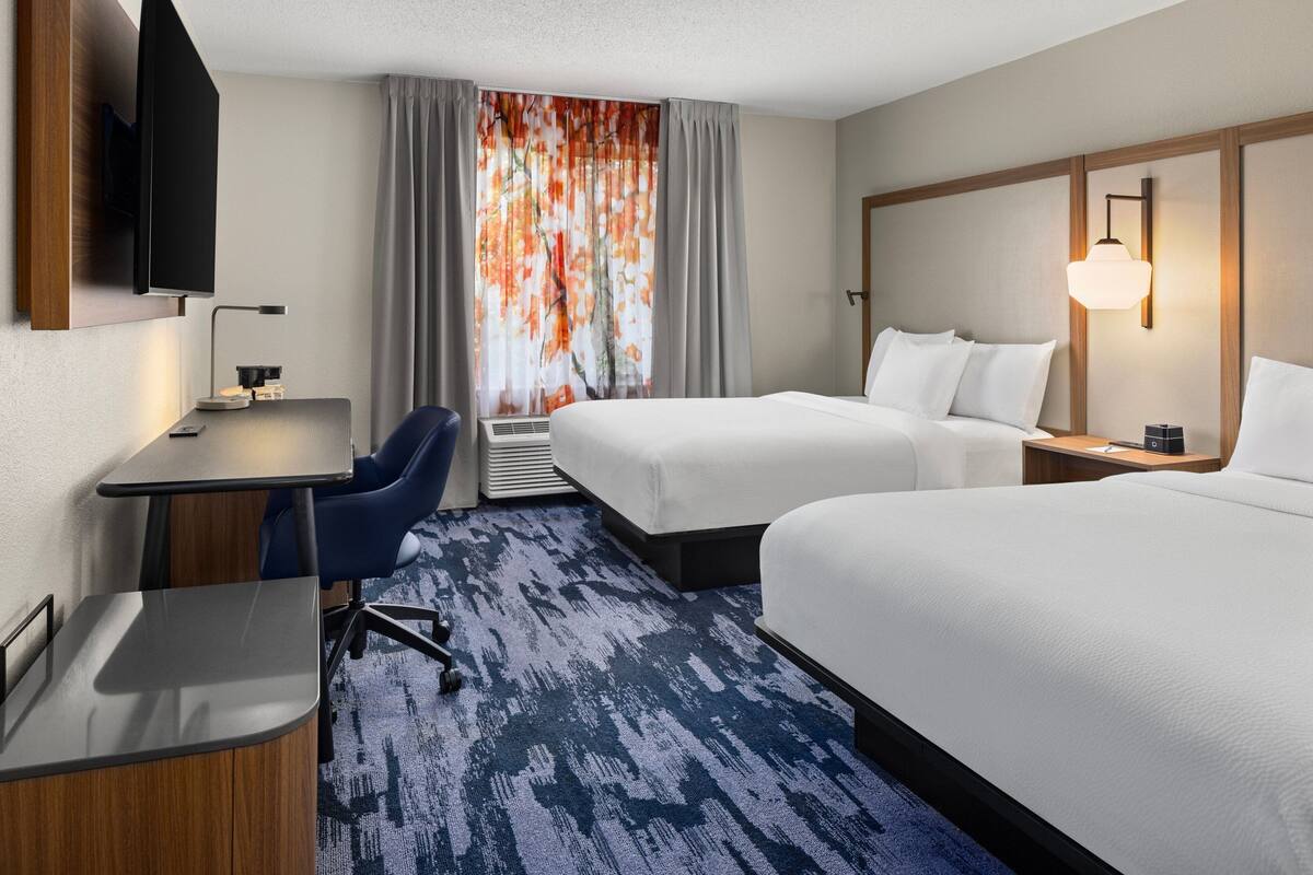 A hotel room with a desk, two beds, a TV, and a large window with curtains.