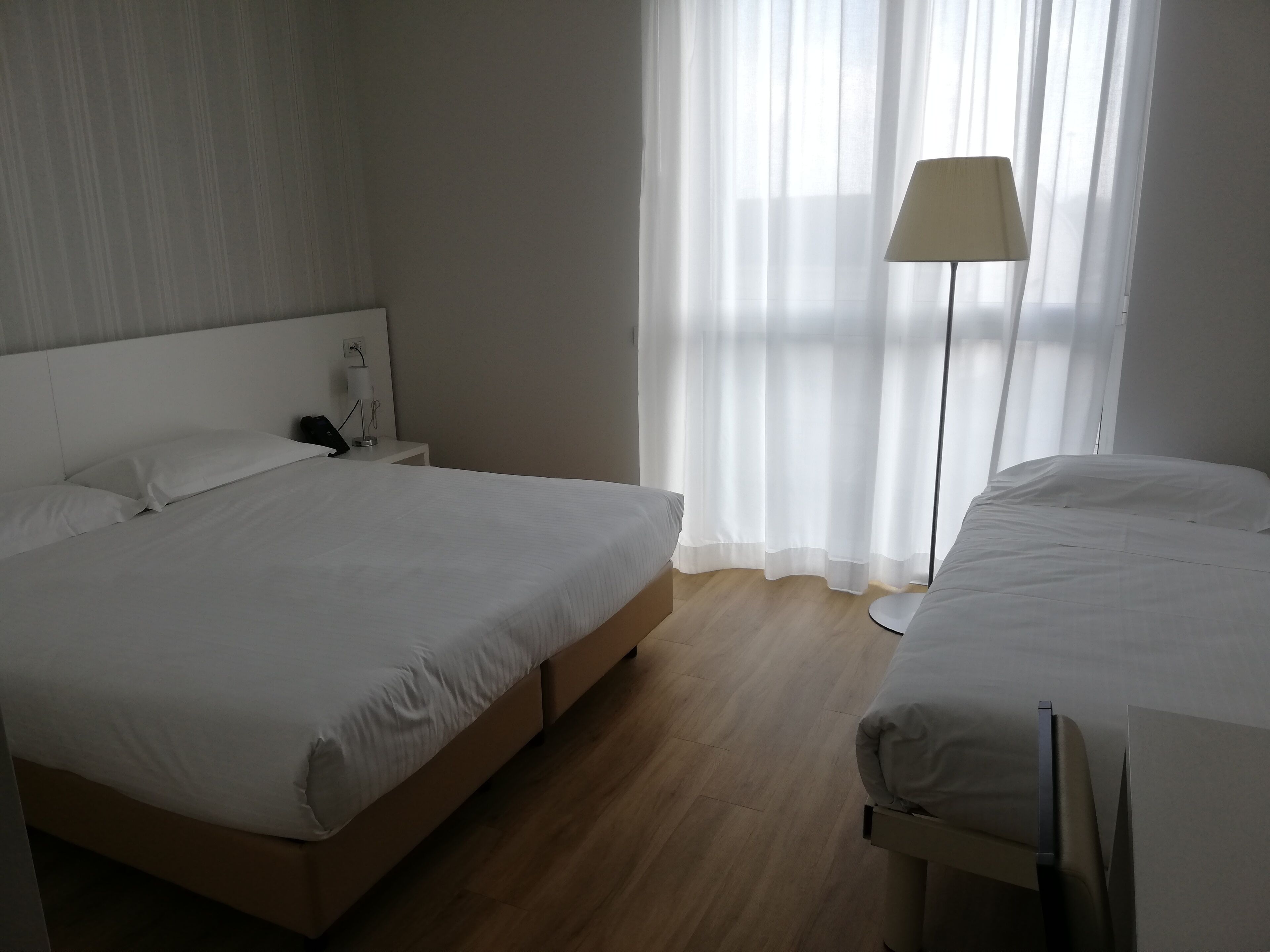Quarto de hotel com cama grande, mesa de cabeceira, luminária e vista para o exterior através de cortinas translúcidas.