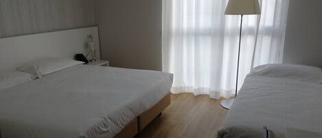 A hotel room with a large bed, bedside table, lamp, and a view of the outdoors through sheer curtains.