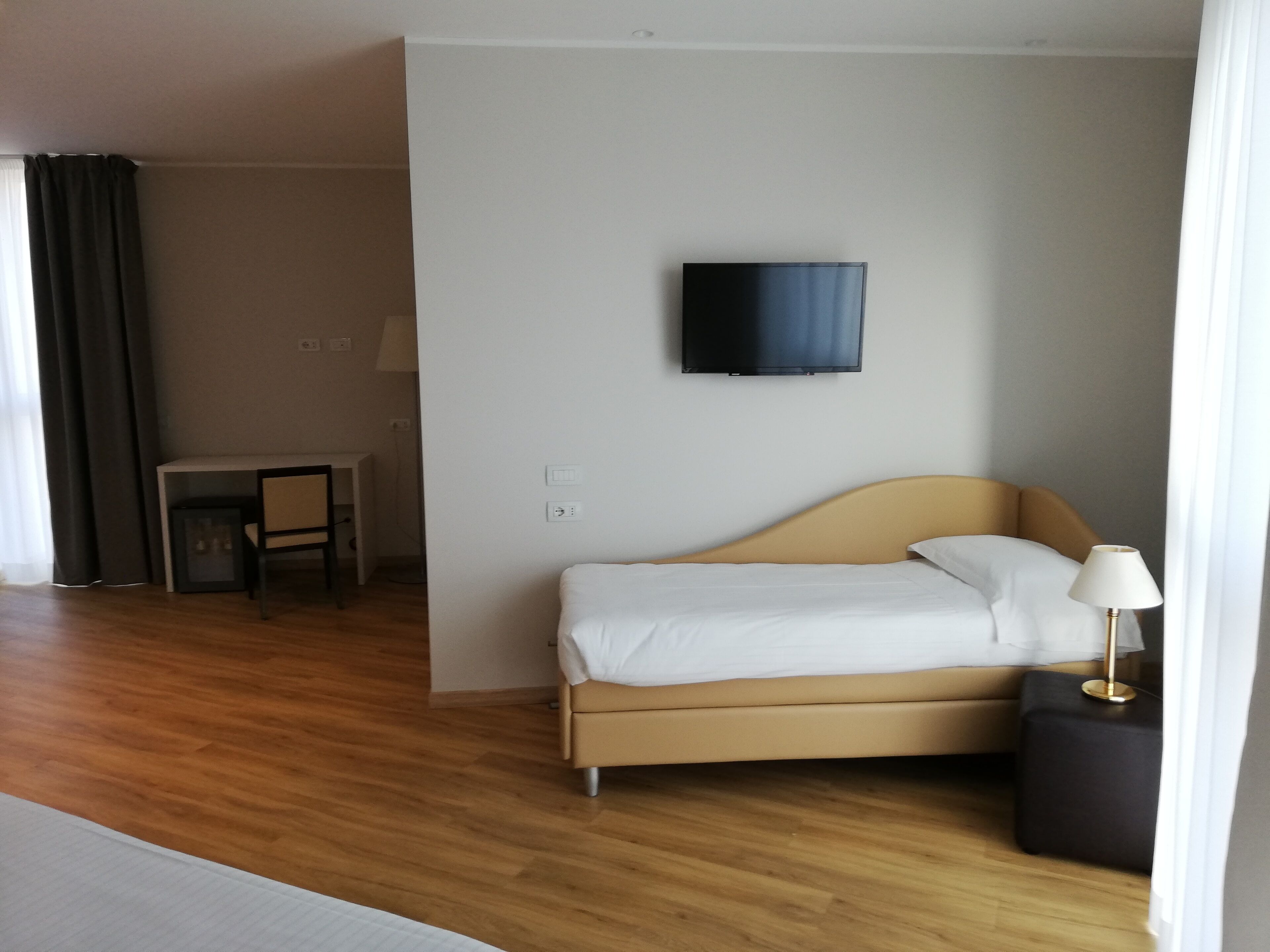 A hotel room with a bed, a TV mounted on the wall, a desk with a chair, and a lamp on a side table.