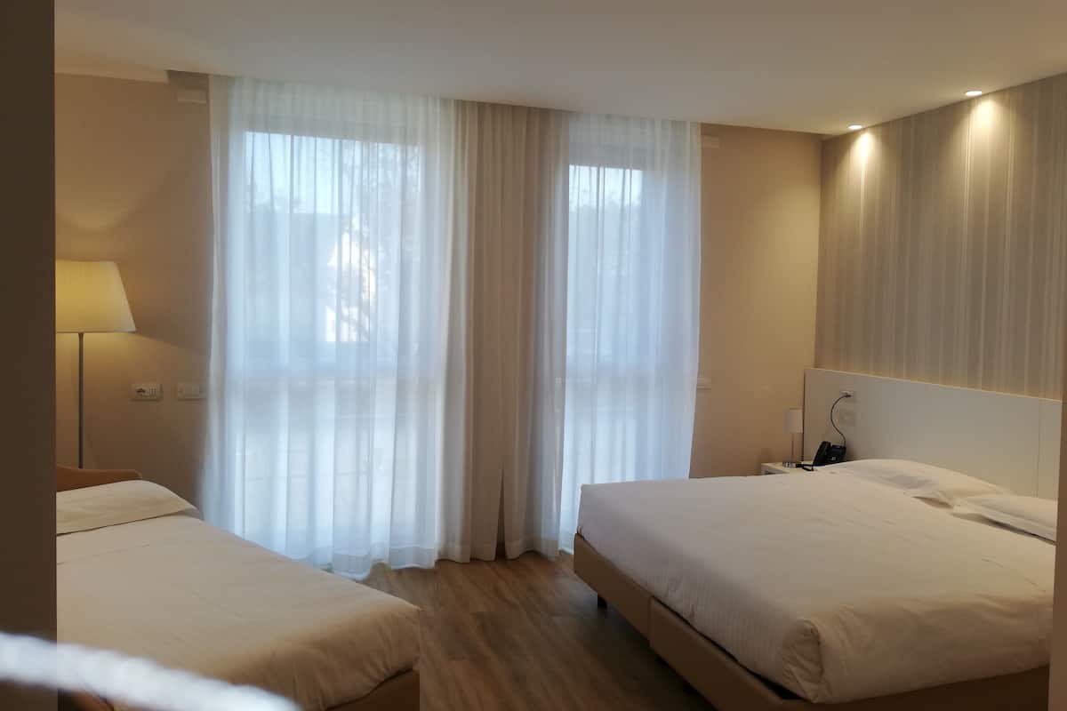 Hotellihuone, jossa on kaksi sänkyä, yöpöytä lampulla ja suuri ikkuna, jossa on ohuet verhot.