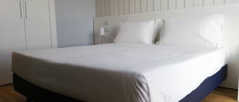 A modern bedroom with a large bed, white bedding, a wooden floor, and a white wardrobe.
