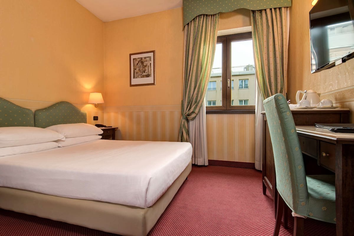 A hotel room with a bed, a desk with a chair, a television, and a window with curtains.