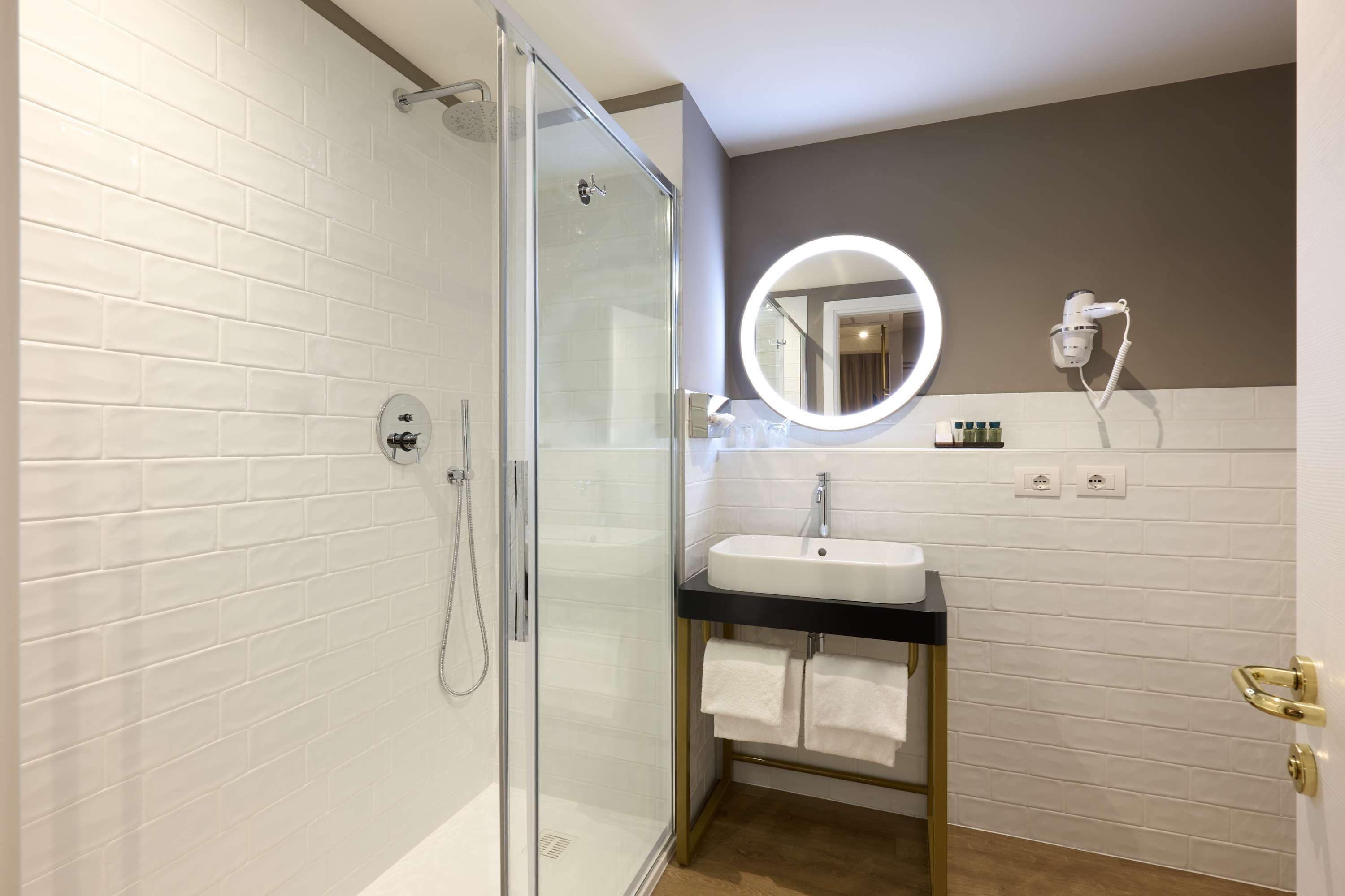 A modern bathroom with a glass-enclosed shower, a white sink with a gold faucet, and a round mirror with a light.