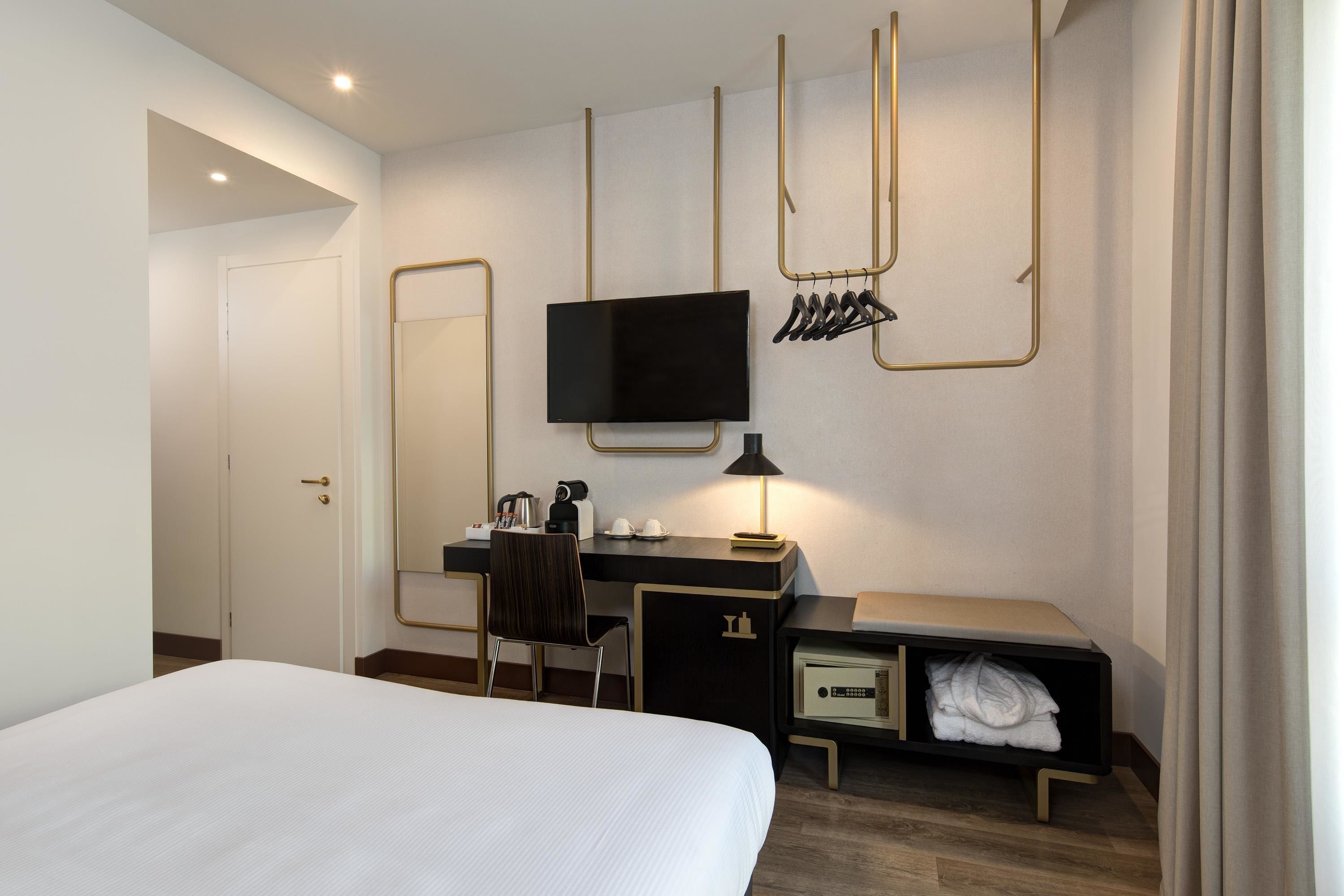 A hotel room with a bed, a desk with a TV, a chair, a desk lamp, a coat rack, and a small cabinet with towels.