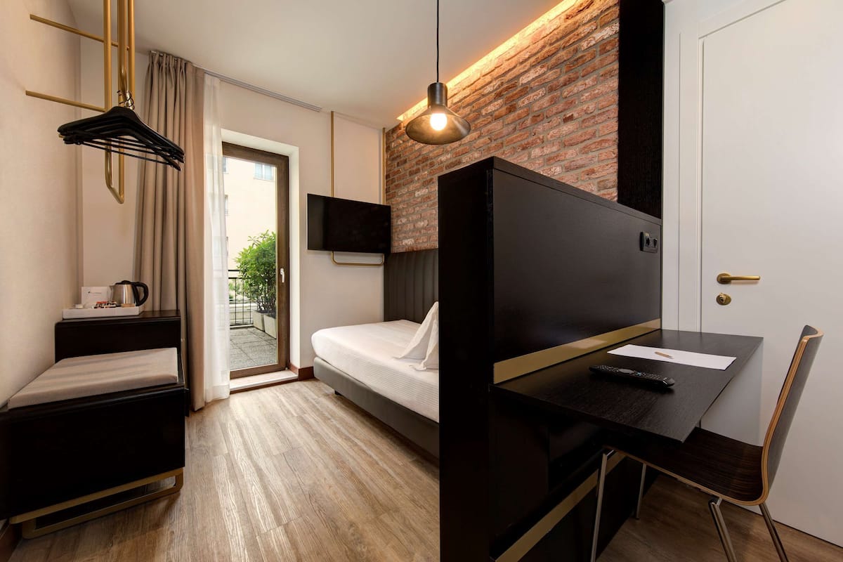 A modern hotel room with a bed, a desk with a chair, a hanging light fixture, and a brick wall.