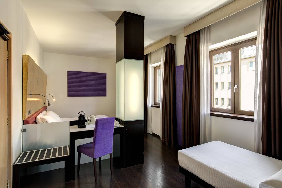 A hotel room with a bed, a desk with a chair, a small kitchenette, and a window with curtains.