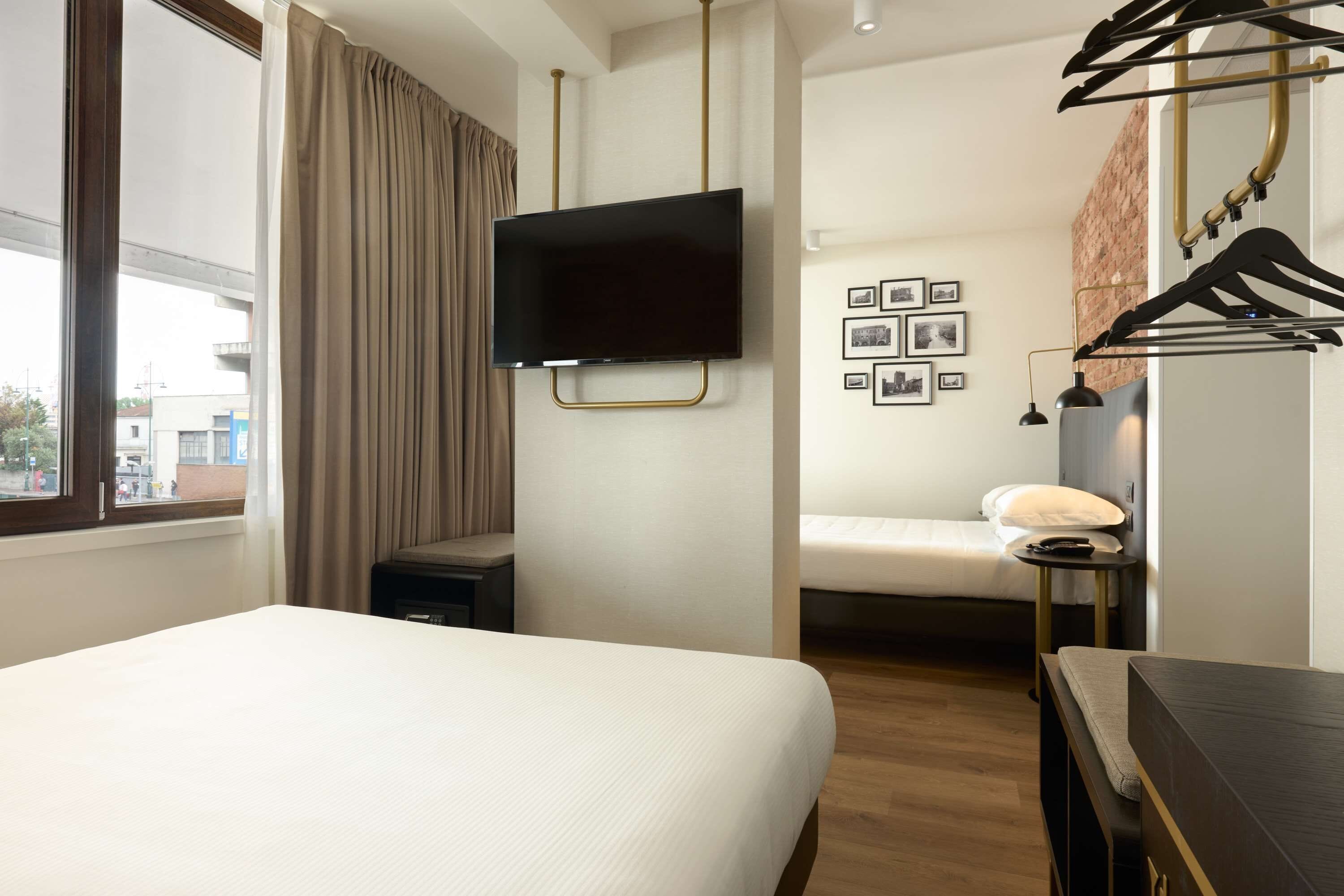 A hotel room with a large bed, a TV mounted on the wall, a bedside table, a small bedside table with a lamp, and a wall-mounted coat rack.