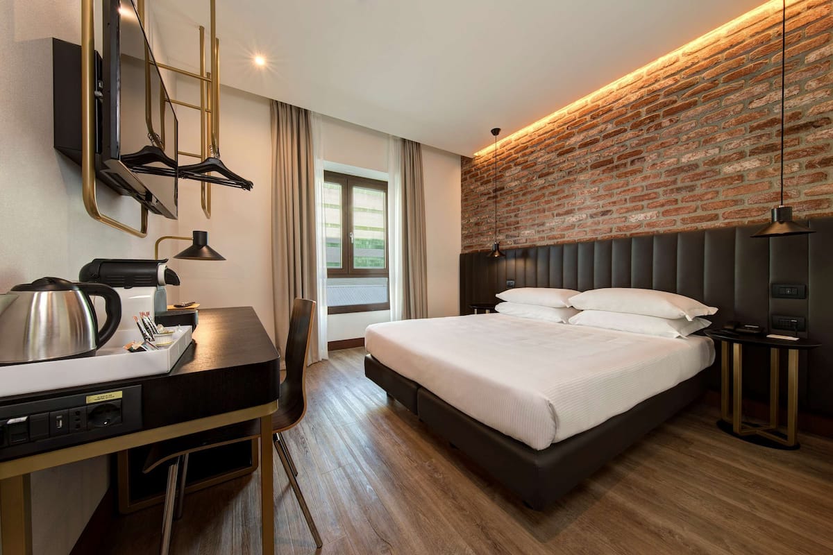 A modern hotel room with a brick accent wall, a large bed, a desk with a kettle, and a TV mounted on the wall.