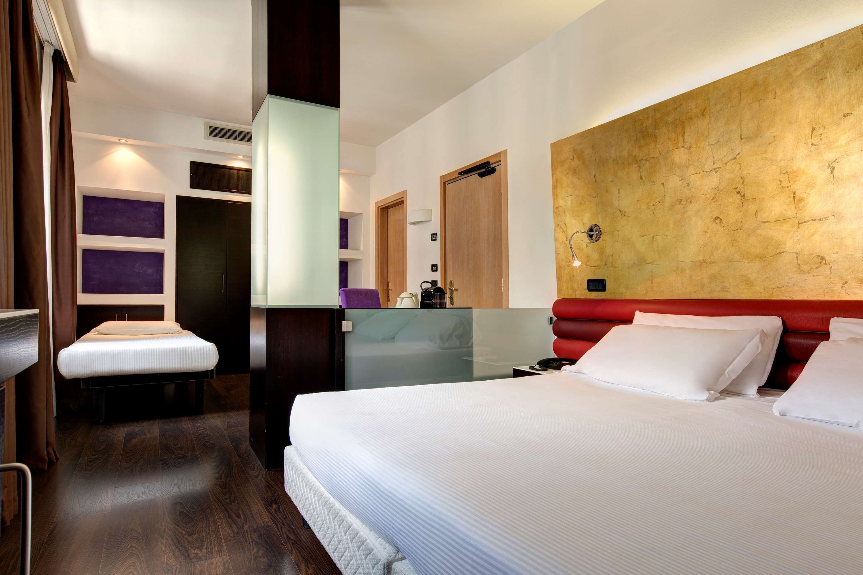 A hotel room with a large bed, a bedside table, a mirror, a wardrobe, and a red headboard.