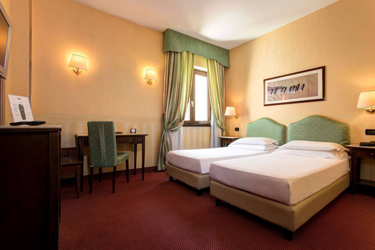 A hotel room with two beds, a desk, a chair, a TV, and a window with curtains.