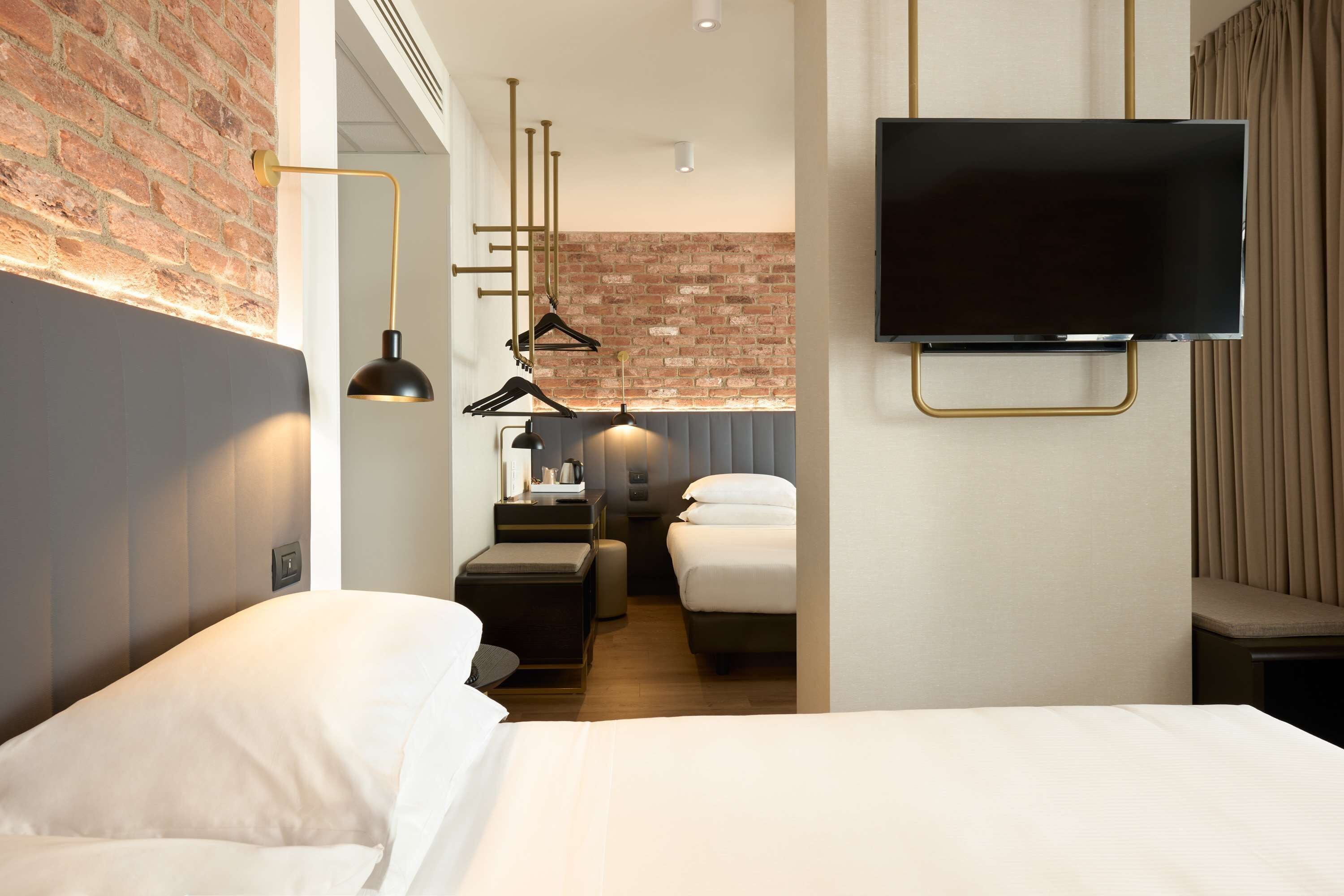 Ein modernes Hotelzimmer mit einer Ziegelfassade, einem Flachbildfernseher, einem Bett mit weißen Bettbezügen, einem Nachttisch und einer Pendelleuchte.