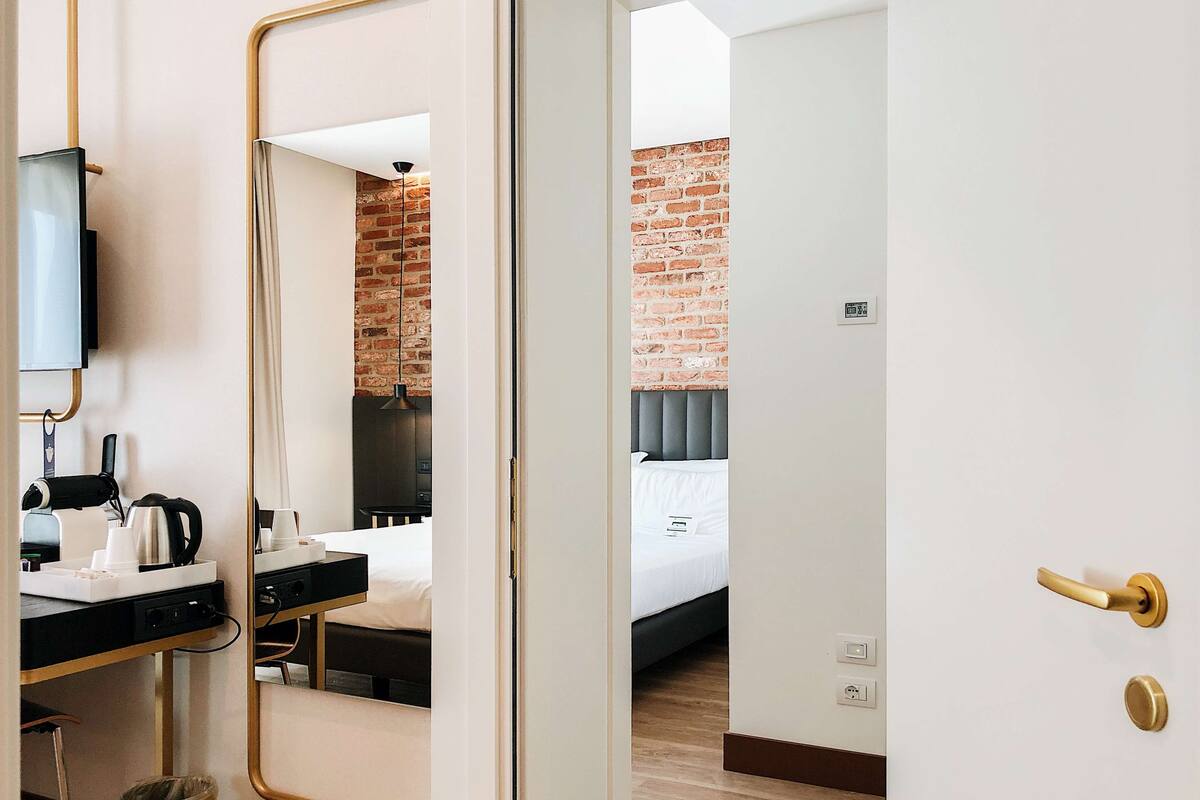 A hotel room with a mirror, a desk with a kettle, a trash bin, a bed with a grey headboard, and a brick wall.
