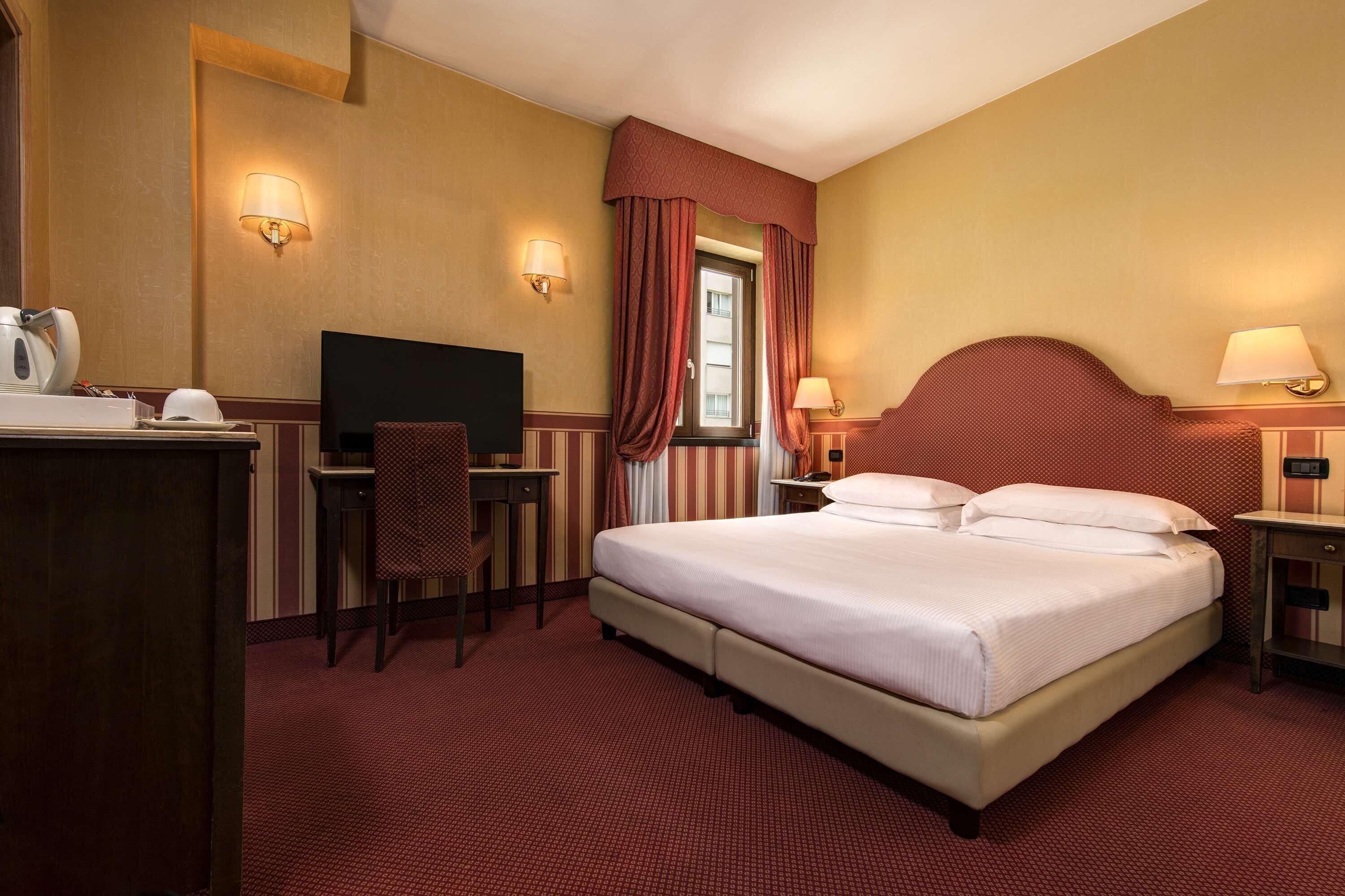 Une chambre d’hôtel équipée d’un lit, d’un bureau, d’une chaise, d’une télévision et d’une fenêtre avec des rideaux.