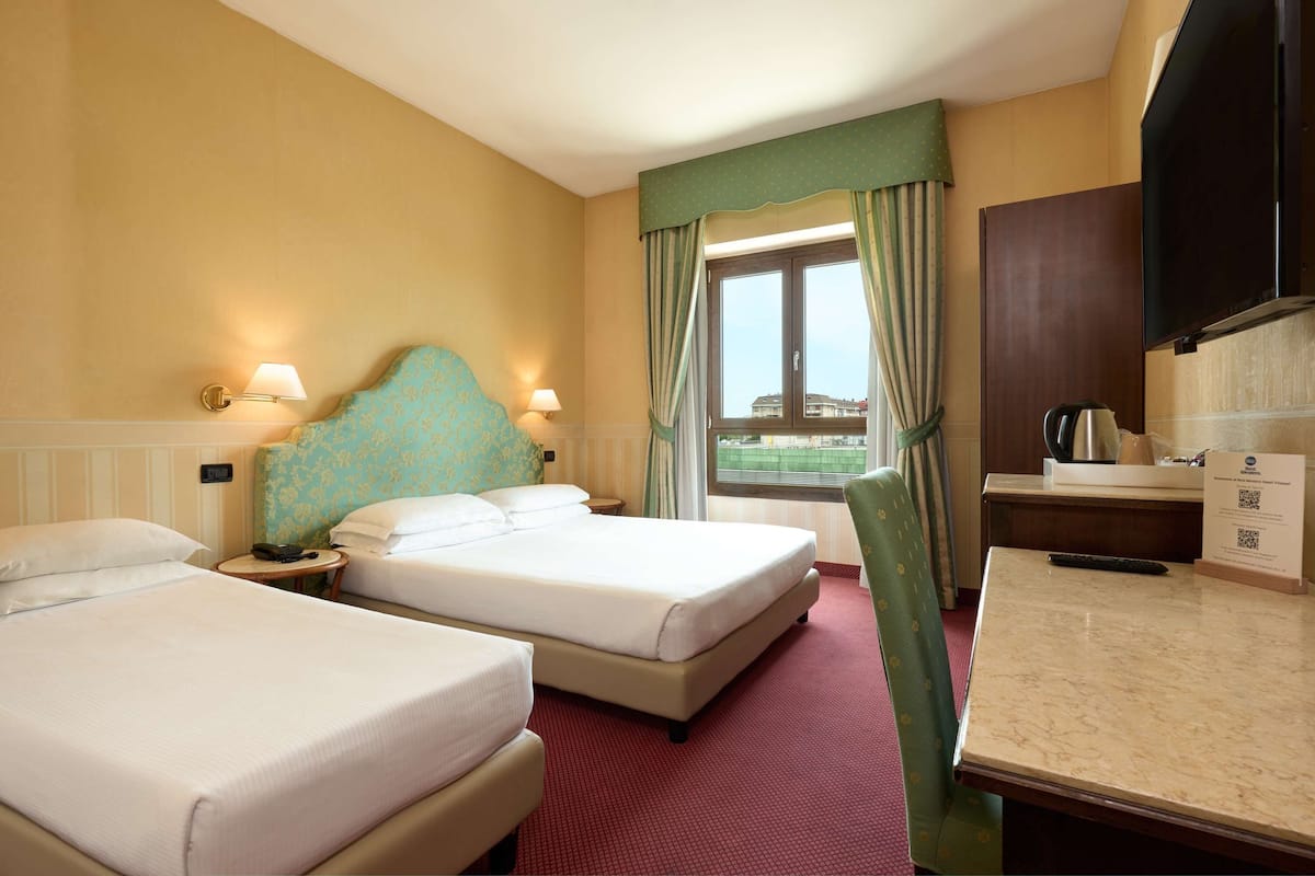 A hotel room with two beds, a desk, a TV, and a window with curtains.