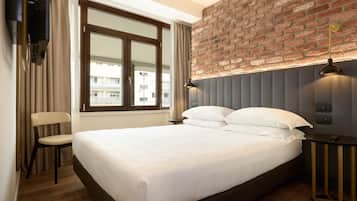 Quarto de hotel com uma cama grande, parede de tijolos, janela com cortinas, uma cadeira e mesa de cabeceira com abajur.