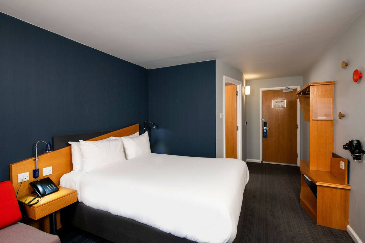 A hotel room with a large bed, a bedside table with a phone, a red chair, and a wooden cabinet.