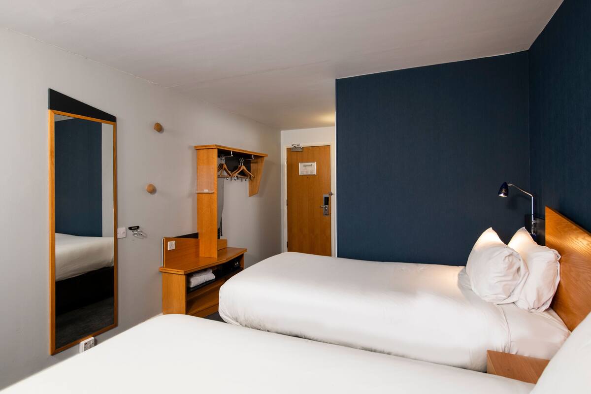 A hotel room with two beds, a wooden desk, a mirror, and a wardrobe.