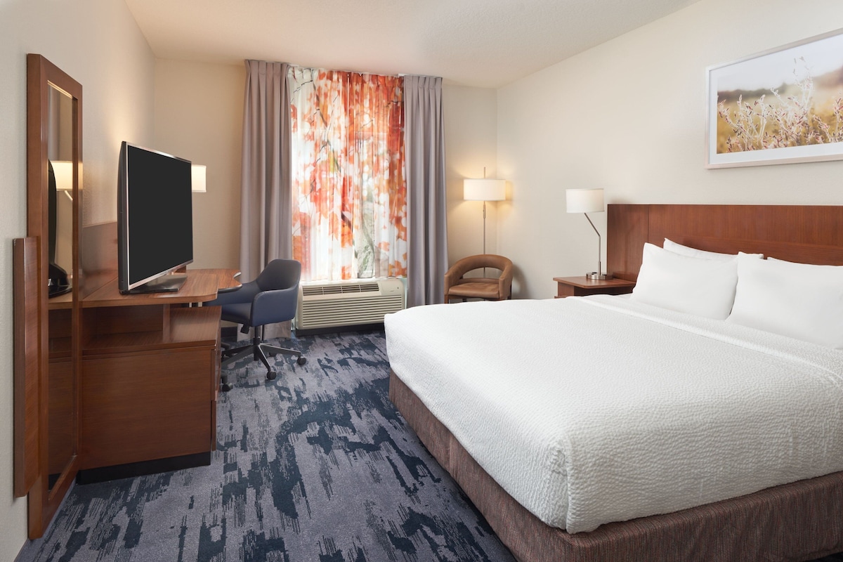 A hotel room with a large bed, a desk with a flat-screen TV, a chair, a desk lamp, and a large window with curtains.