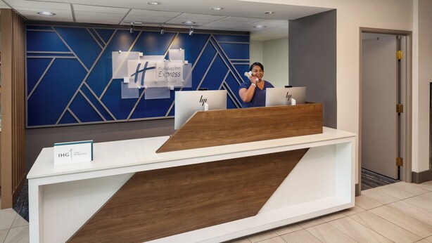 Ein Empfangsschalter mit modernem Design, in Weiß und Holz gehalten. An der Wand hinter dem Schalter befinden sich Schilder für Holiday Inn Express und IHG.
