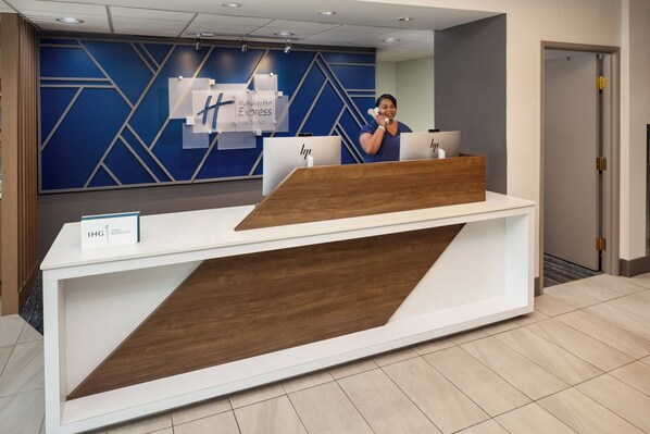 Un comptoir d’accueil au design moderne, avec une finition blanche et en bois. Des panneaux indiquant « Holiday Inn Express » et « IHG » sont accrochés au mur derrière le comptoir.