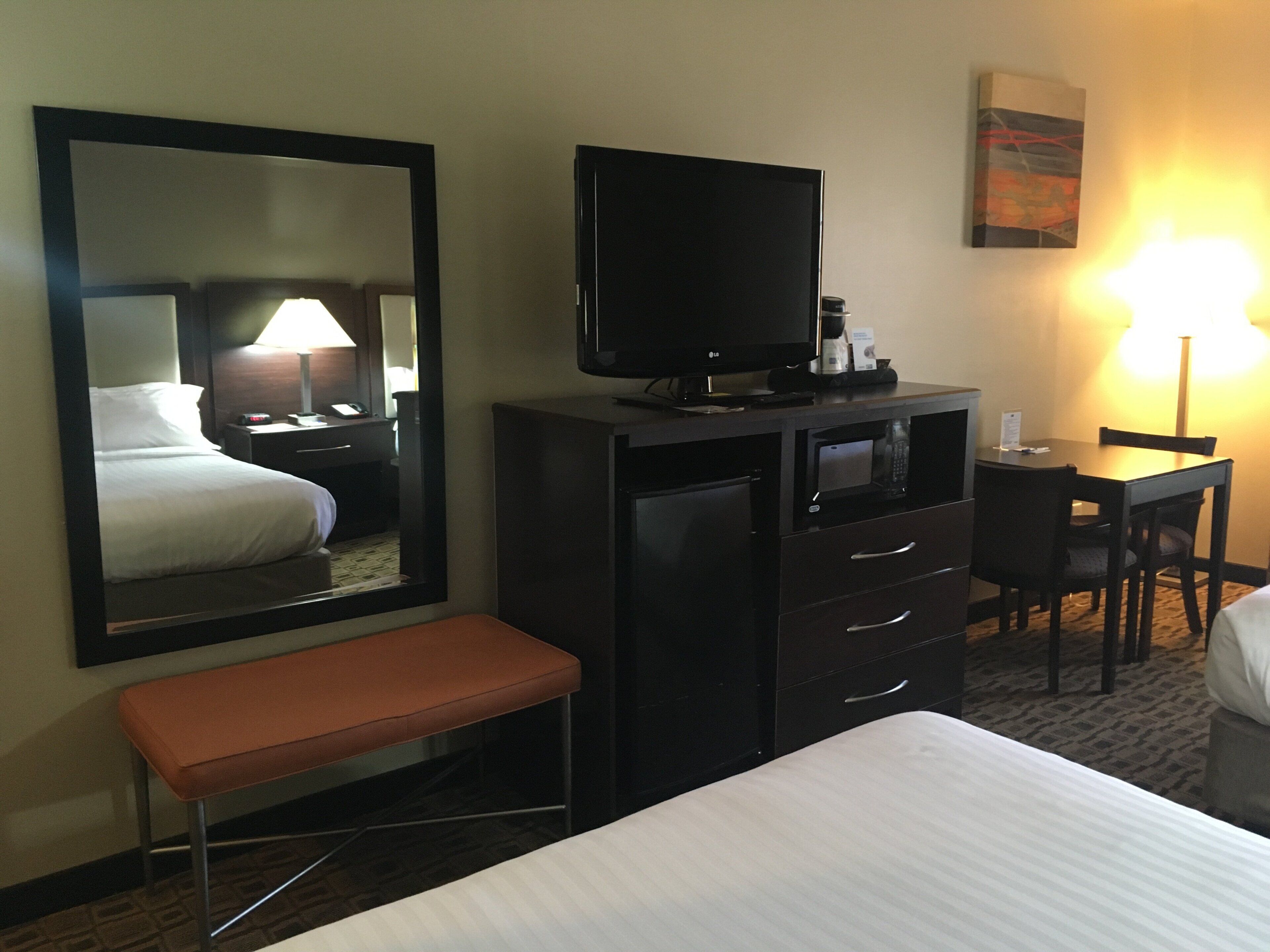 A hotel room with a bed, a desk, a television, a mirror, and a bench.